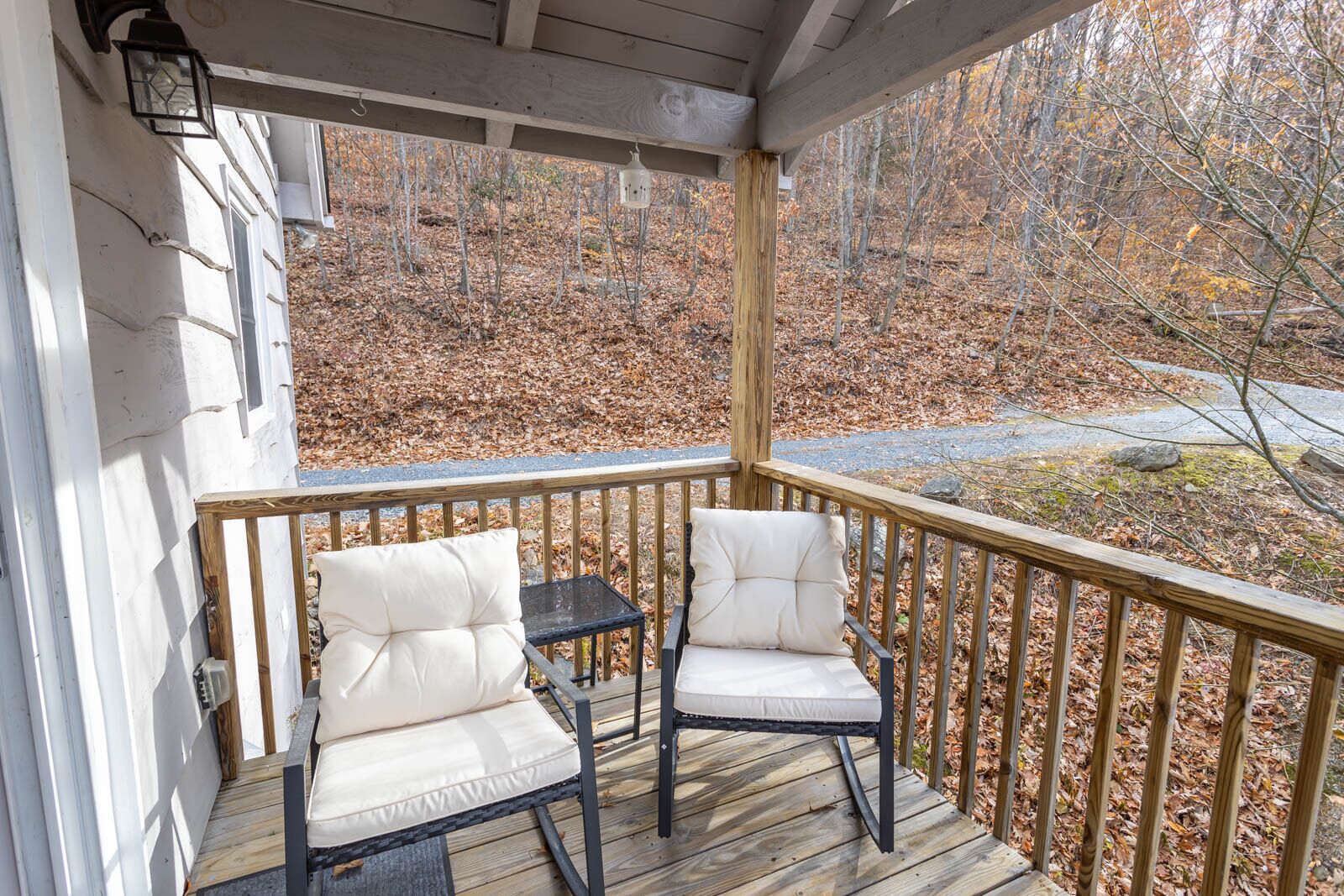 A covered porch with two cushioned chairs and a small table, overlooking a wooded area.
