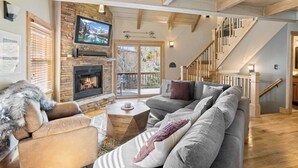 Un salon chaleureux avec une cheminée, un grand canapé d’angle gris, un mur décoratif en pierre et un escalier en bois.