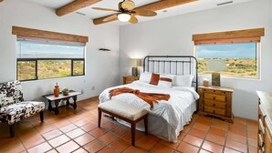 A bedroom with a bed, a chair, a bench, a ceiling fan, and two windows with a view. - Stunning Views, Pool, Sauna, Steam Shower, Firepit! (Cottonwood)