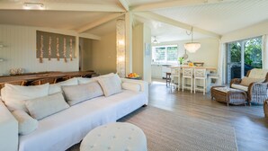 A spacious living room with a white sofa, a dining area with a wooden table, and a kitchen with white cabinets. - Orion Sands: Absolute beachfront with private steps to beach (Vincentia)