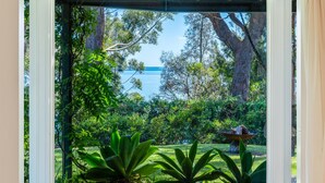 A view through a window with a garden, trees, and a body of water. - Orion Sands: Absolute beachfront with private steps to beach (Vincentia)