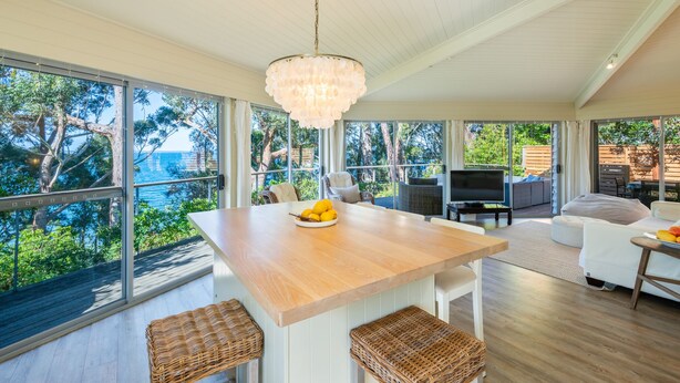 A modern living room with a wooden table, wicker stools, a sofa, and a TV. Large windows offer a view of trees and the sea. - Orion Sands: Absolute beachfront with private steps to beach (Vincentia)