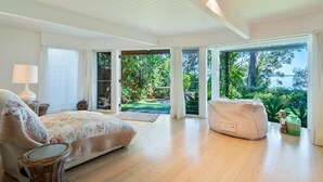 A spacious room with a large bed, a chair, and a bean bag. It has a view of a garden and a body of water through large windows. - Orion Sands: Absolute beachfront with private steps to beach (Vincentia)