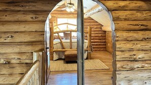A log cabin interior with a central hallway leading to a bedroom with a bed and a window.