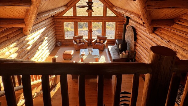 A log cabin interior with a central seating area, a fireplace, and a view through the large windows.