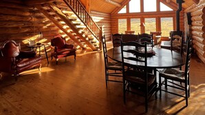A log cabin interior with a dining area, wooden floors, a staircase, and a large window.