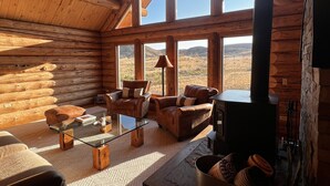 A log cabin interior with a fireplace, leather sofas, a glass coffee table, and large windows offering a view of the desert landscape.
