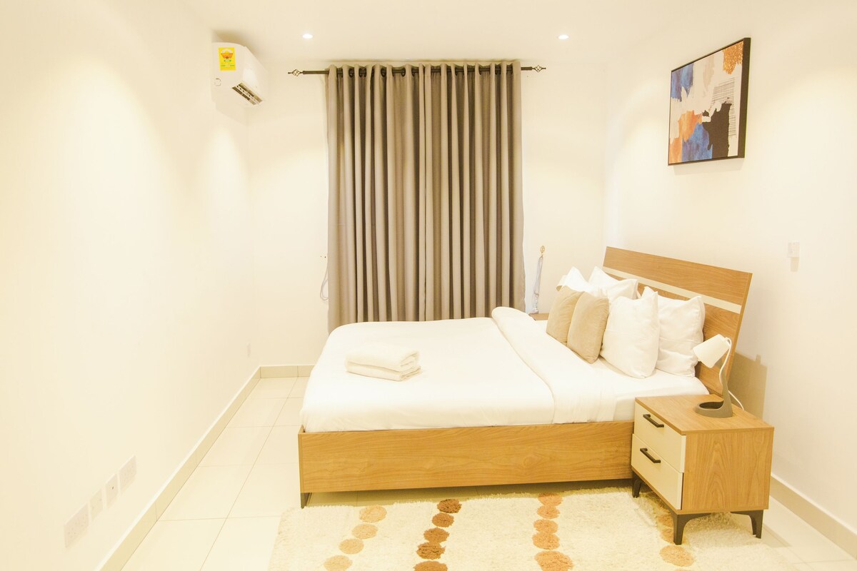 Een hotelkamer met een houten bed, een nachtkastje, een wandairconditioner, een raam met gordijnen en een schilderij aan de muur.