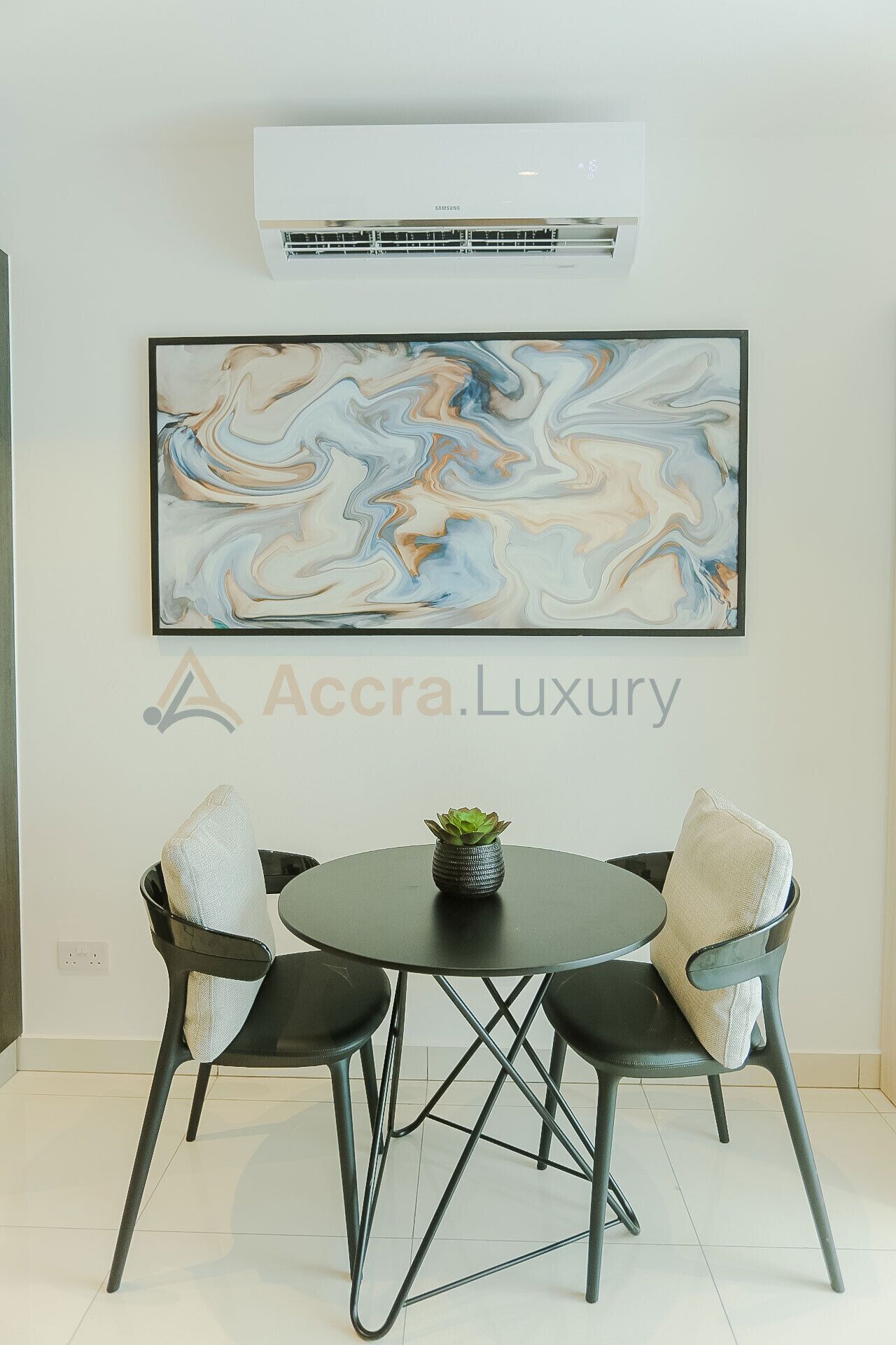 A modern dining area with a round table, two chairs, a potted plant, and a large abstract painting on the wall.