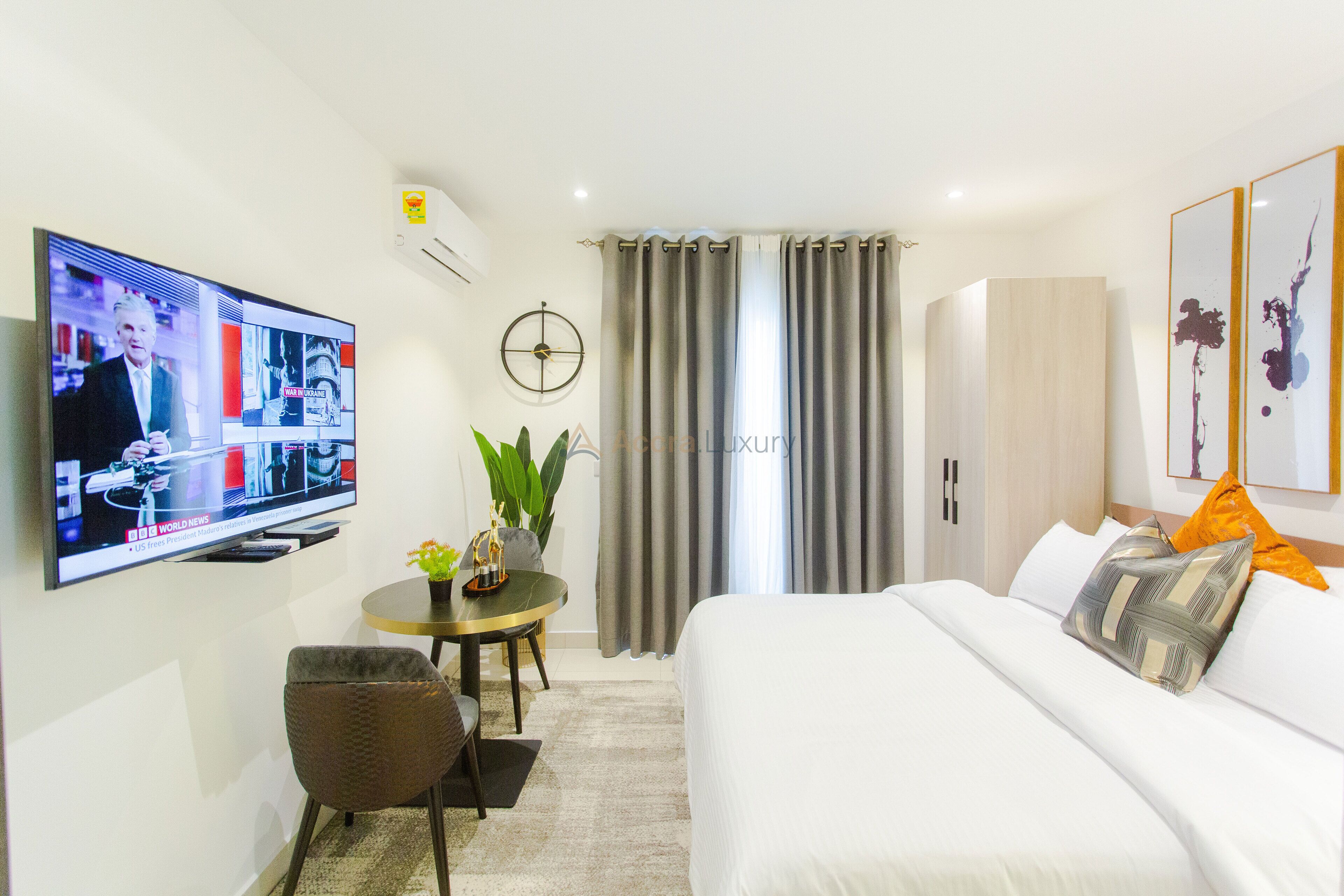 A hotel room with a bed, a TV mounted on the wall, a small table with a plant, and a window with curtains.