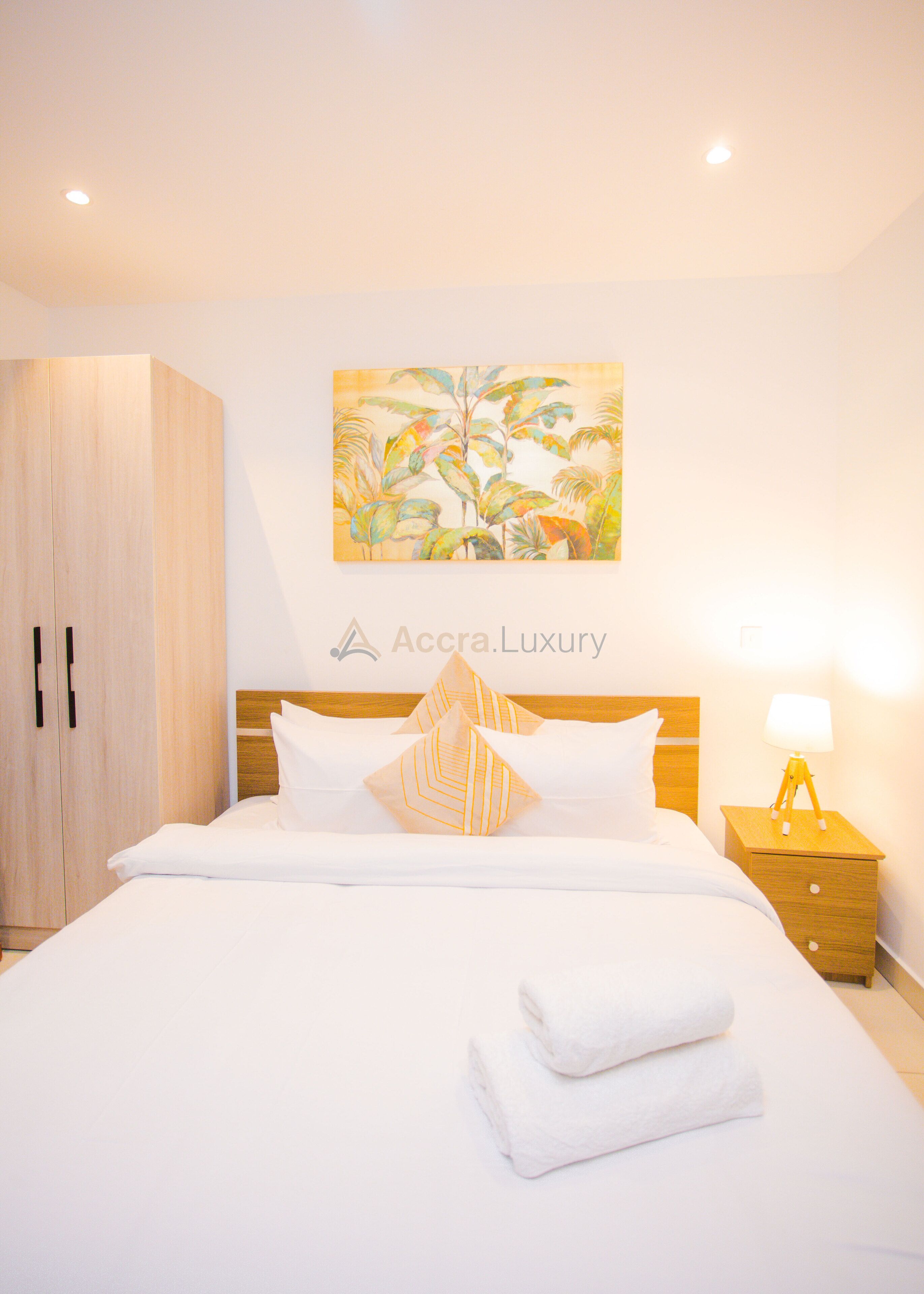 A hotel room with a bed, a nightstand, a lamp, a wardrobe, and a tropical-themed painting on the wall.