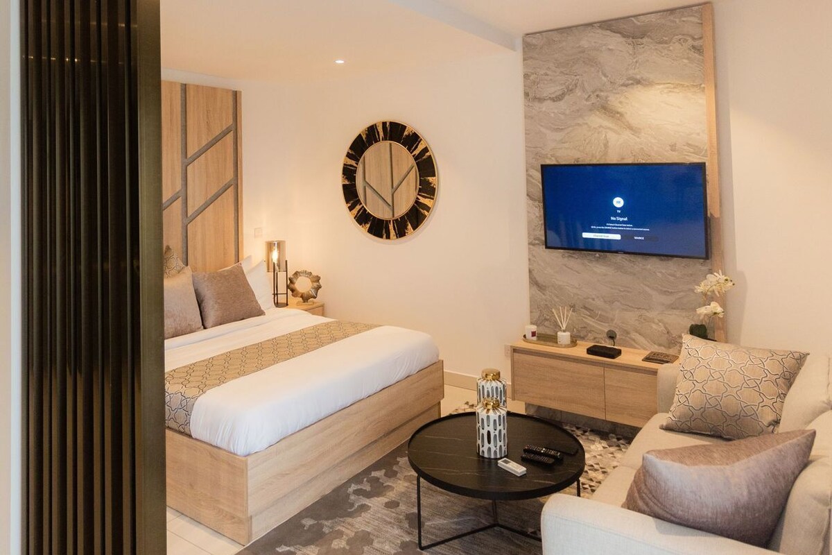 Quarto de hotel moderno com cama, sofá, mesa de centro, TV na parede e um objeto decorativo na parede.