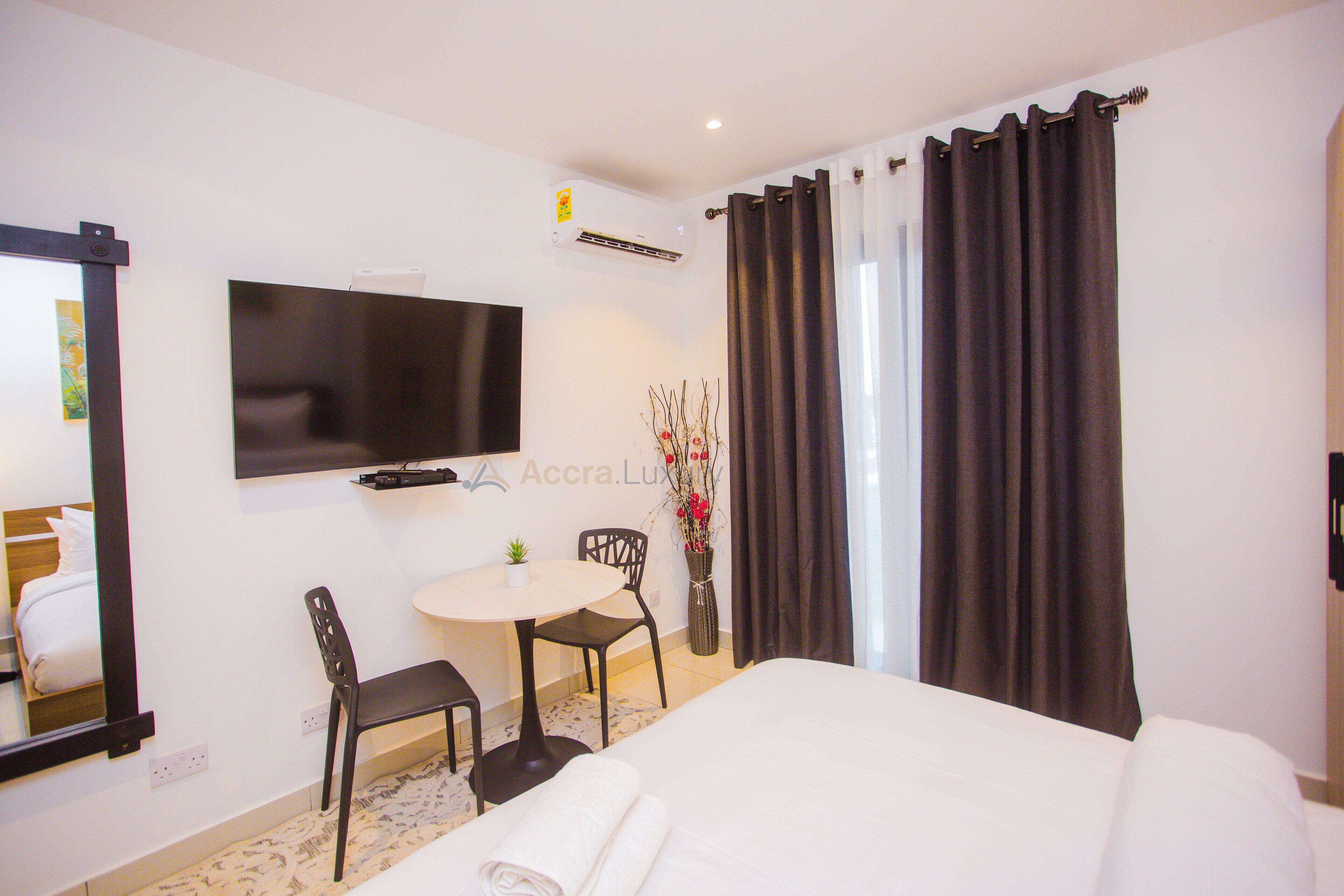 A hotel room with a bed, a TV mounted on the wall, a small table with a chair, and a vase with flowers.