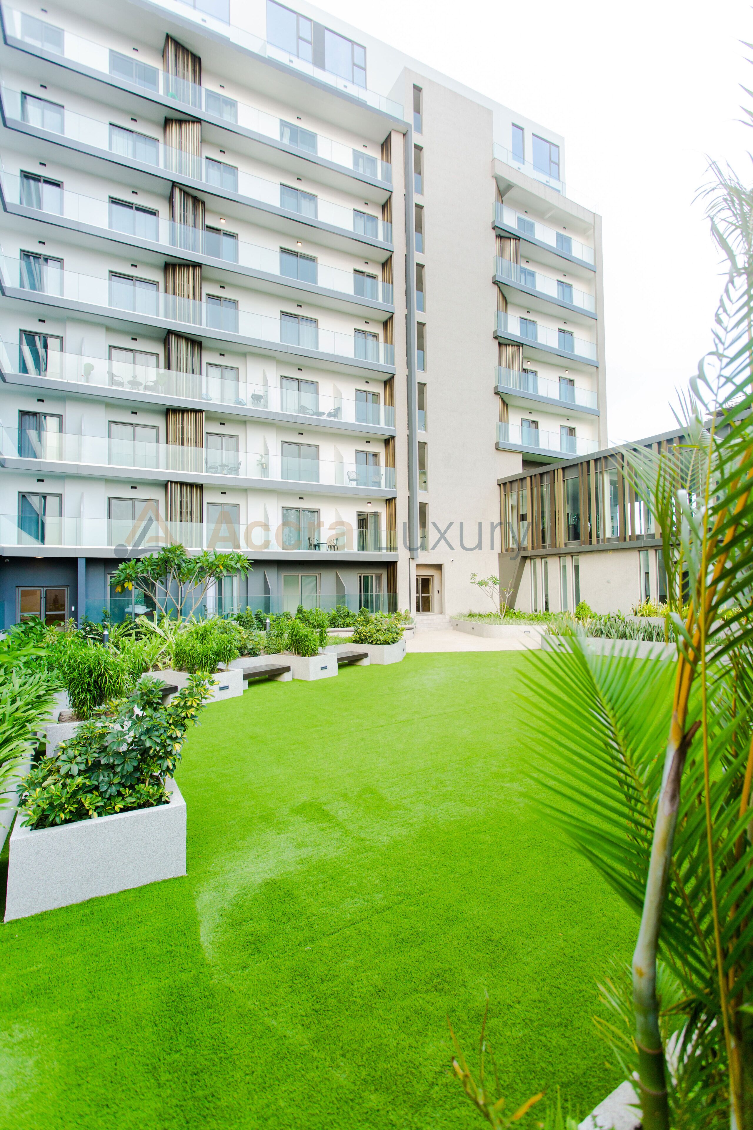 Un immeuble moderne à plusieurs étages, doté de balcons, d’une cour intérieure avec une pelouse artificielle et d’une variété de plantes.