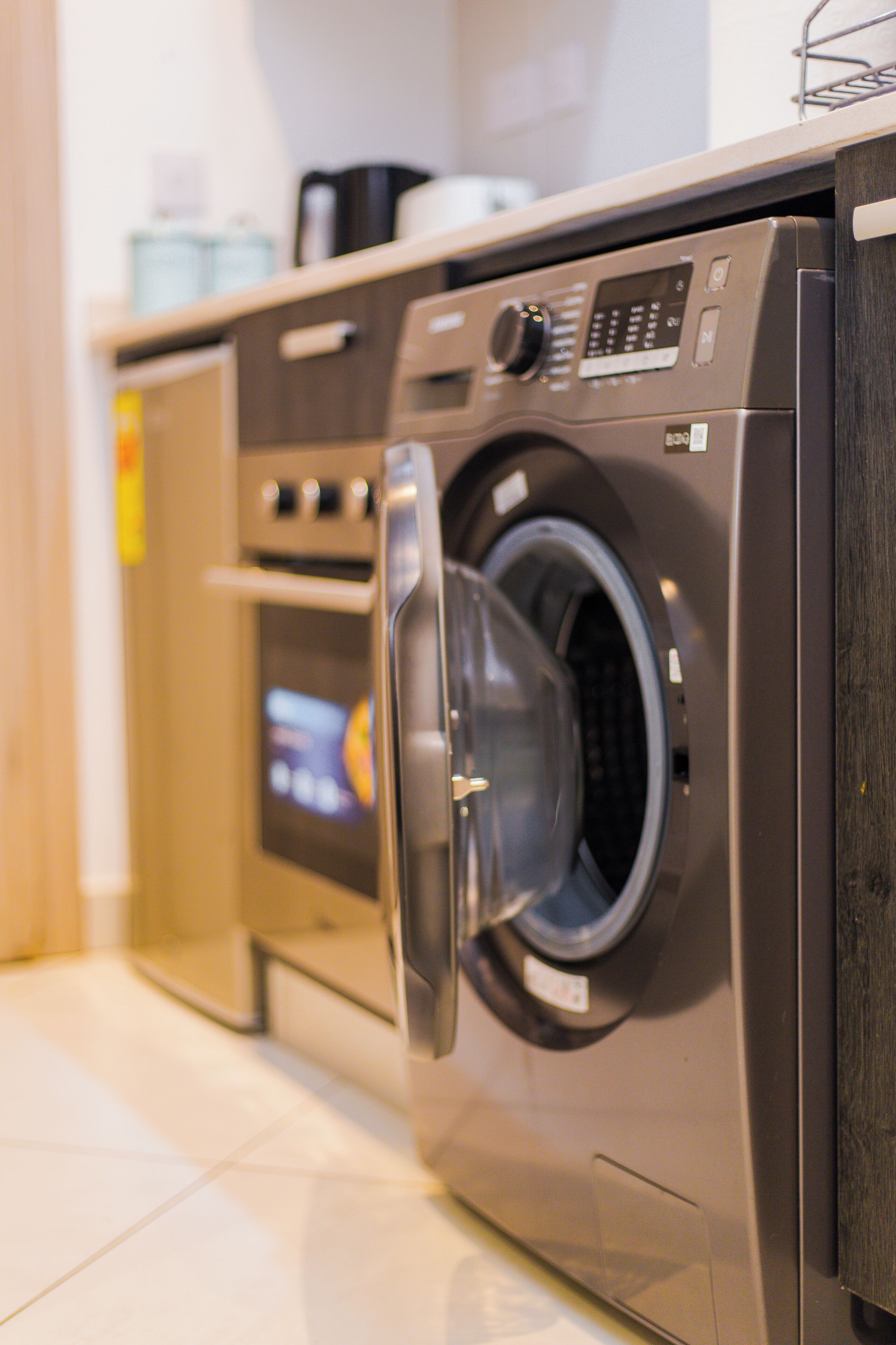 Vue de face d’un lave-linge à chargement frontal moderne, en acier inoxydable, doté d’un écran numérique, placé à côté d’un four encastré et d’une petite machine à café, le tout sur un plan de travail.