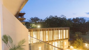 A modern building with a glass facade, a balcony with chairs and plants, and a well-lit outdoor area with a pool.