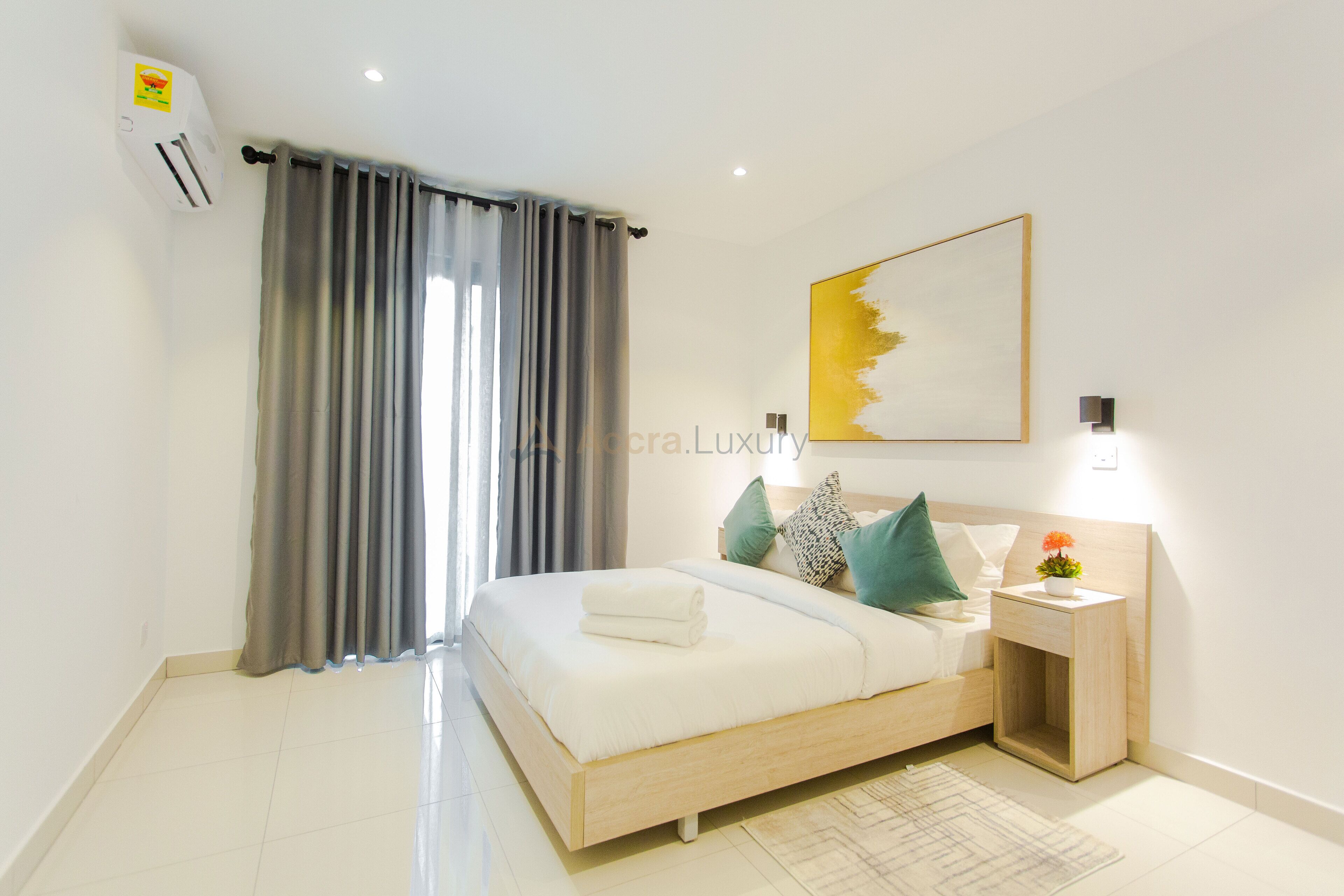 Un dormitorio moderno con cama, cortinas, una obra de arte en la pared y una mesita de noche con lámpara.
