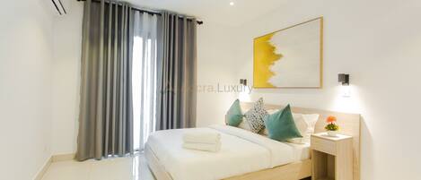 Un dormitorio moderno con cama, cortinas, una obra de arte en la pared y una mesita de noche con lámpara.
