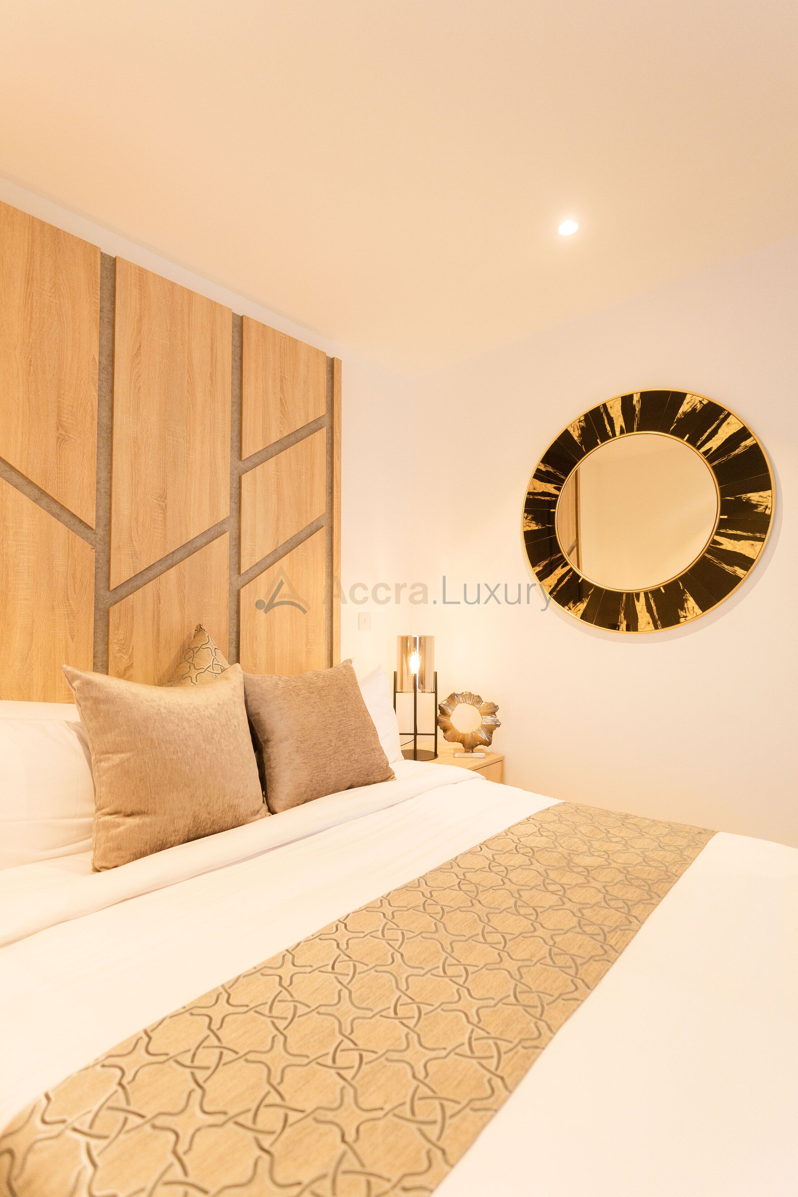A hotel room with a bed, a wooden headboard, a round mirror, and a patterned throw.