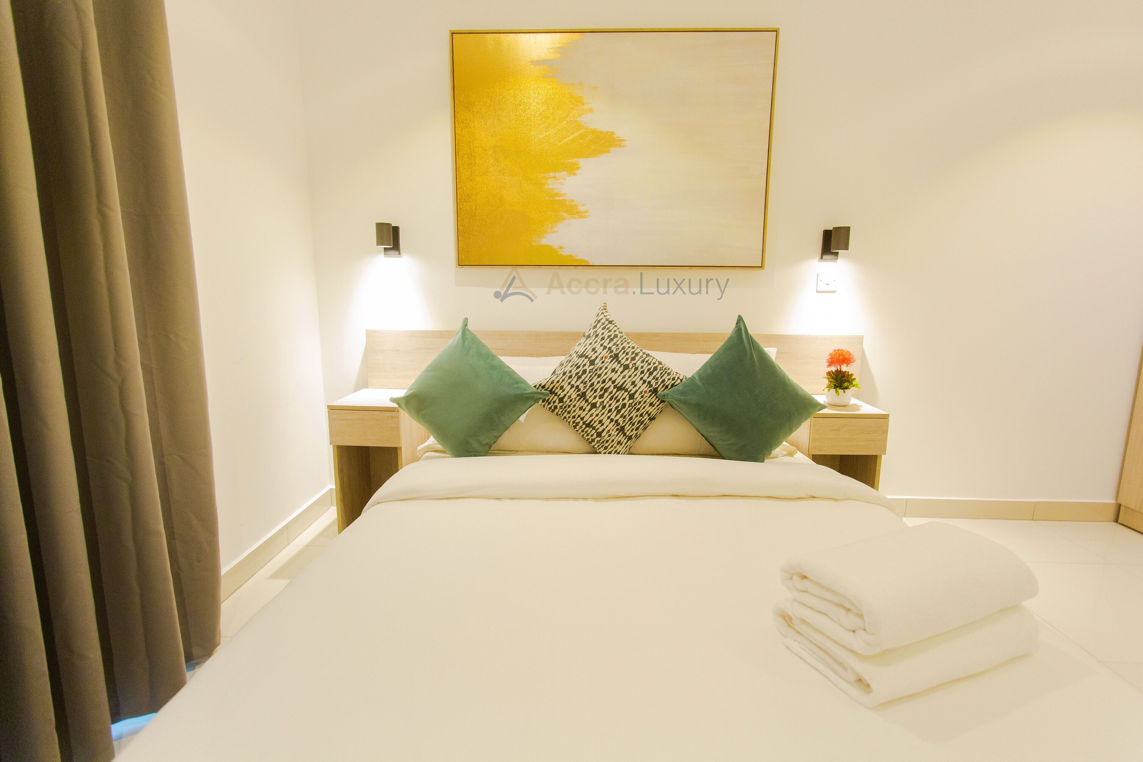 A hotel room with a bed, two bedside lamps, a nightstand with a flower, and a painting on the wall.