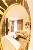 A hotel room with a bed, a sofa, a coffee table, a dining area with chairs, and a large mirror reflecting the room.
