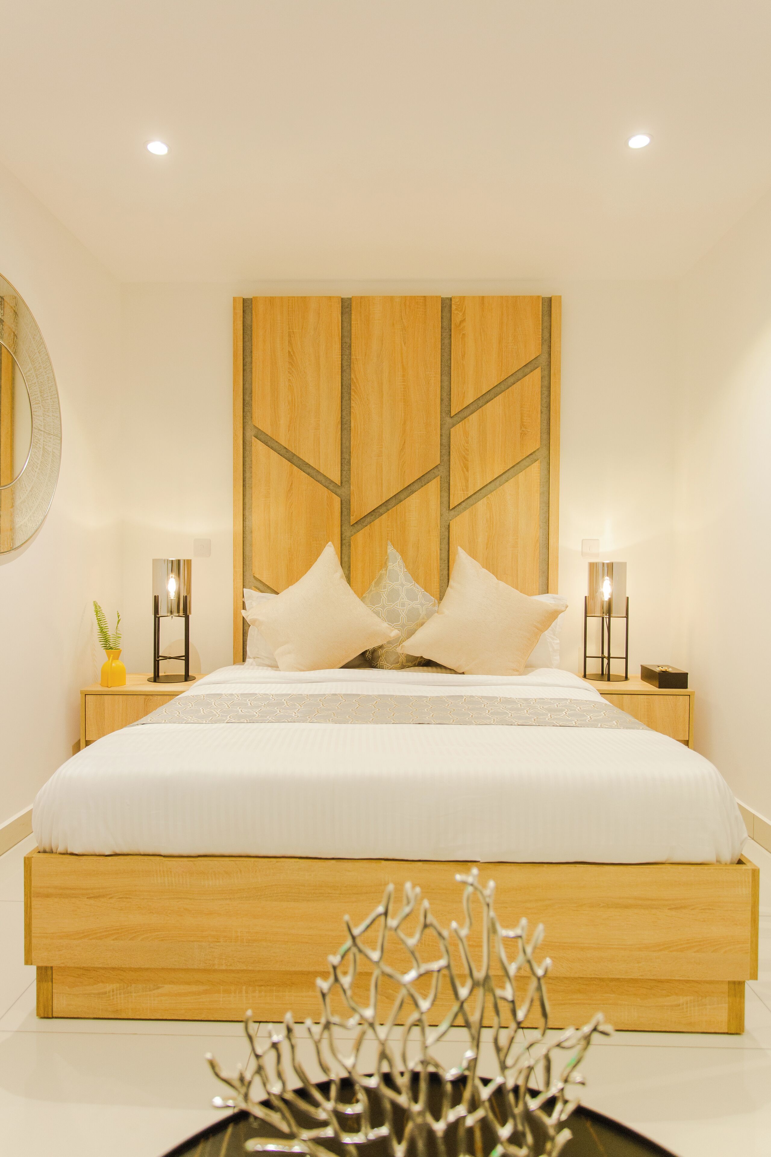 Un dormitorio moderno con cabecera de madera, una cama grande con ropa blanca de cama, dos mesitas de noche con lámparas y una planta decorativa.