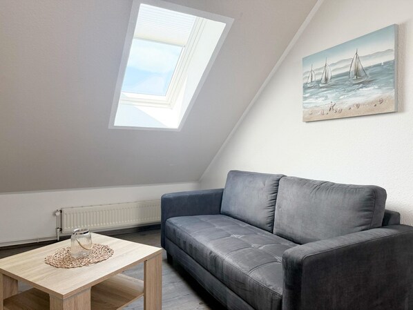 A room with a sloped ceiling, a grey sofa, a wooden table with a glass jar, a radiator, and a painting of sailboats.