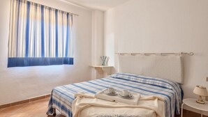 Ein Schlafzimmer mit einem Bett, einem Fenster mit Vorhängen, einem Nachttisch und einer Lampe.