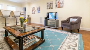 A living room with a glass coffee table, a grey armchair, a flat-screen TV, and framed pictures on the wall.