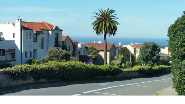 A coastal street with white buildings, a palm tree, and a clear view of the ocean.
