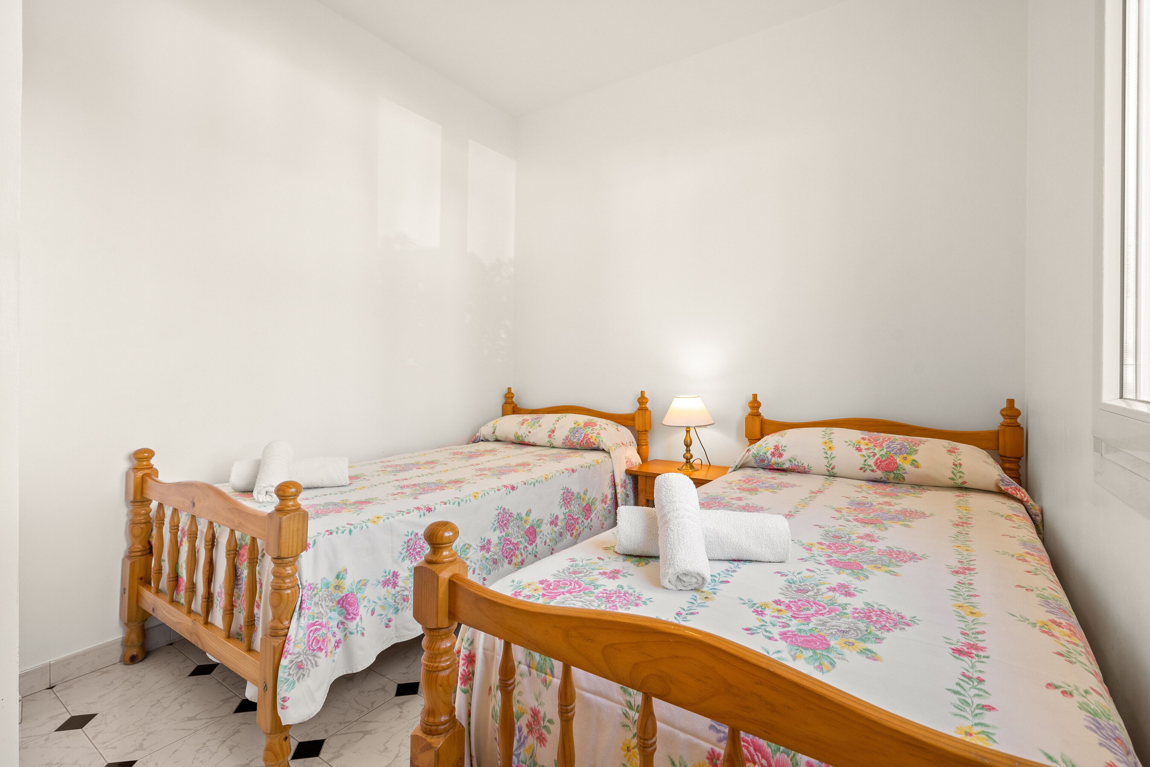 A bedroom with two wooden beds, floral bedding, a bedside table with a lamp, and a window.