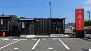 A hotel entrance with a red sign, two black container buildings, and a parking lot with white lines. - HOTEL R9 The Yard Shikokuchuo (Shikokuchuo)