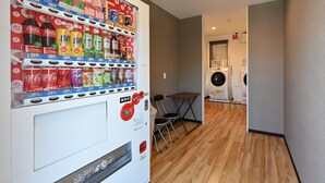 A vending machine stocked with beverages and snacks, a washing machine, and a small table with chairs in a hallway. - HOTEL R9 The Yard Shikokuchuo (Shikokuchuo)
