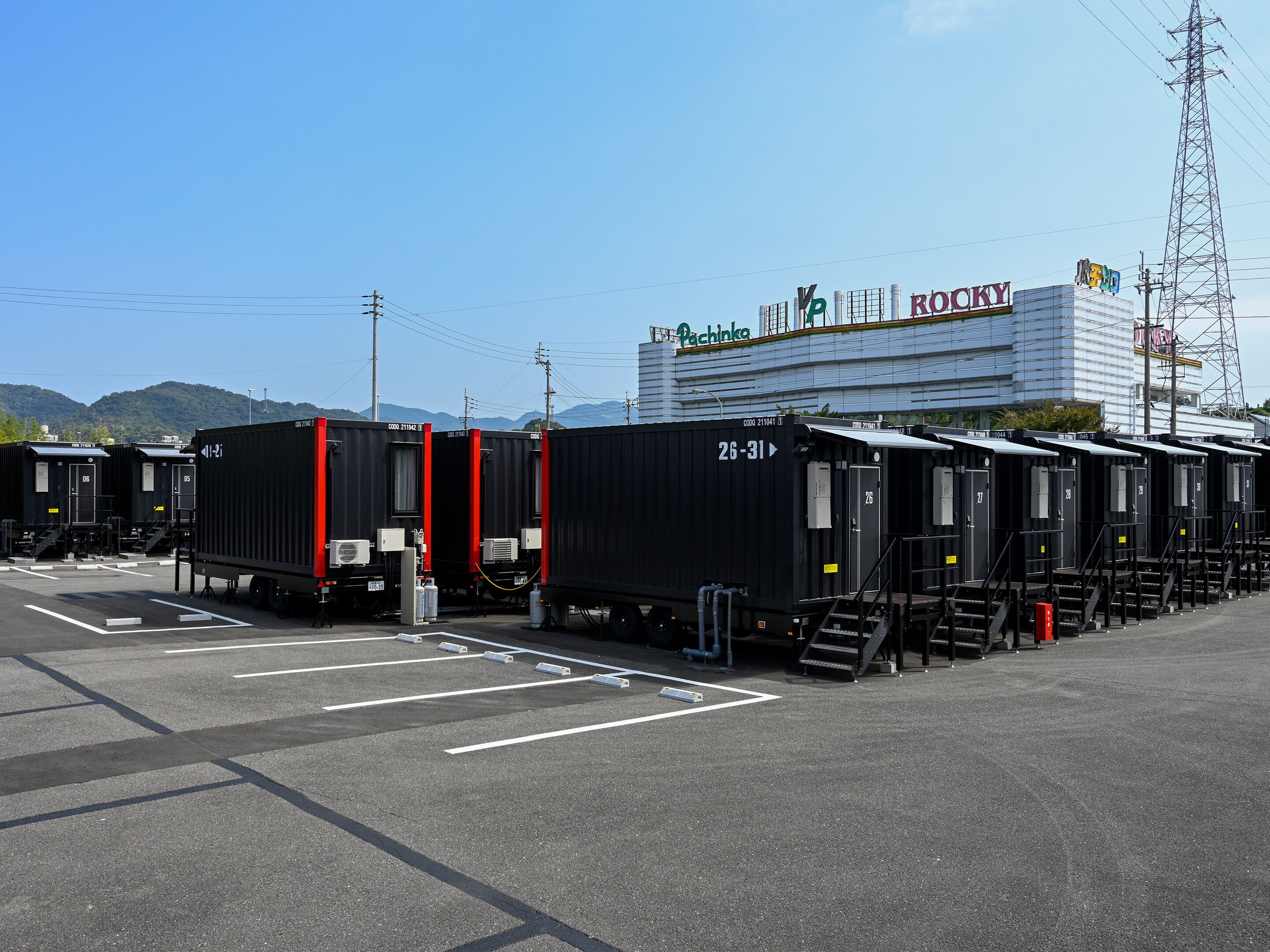 A row of black portable cabins with red accents, numbered 26-31, parked in a lot with a building in the background labeled "Pachinko" and "ROCKY".