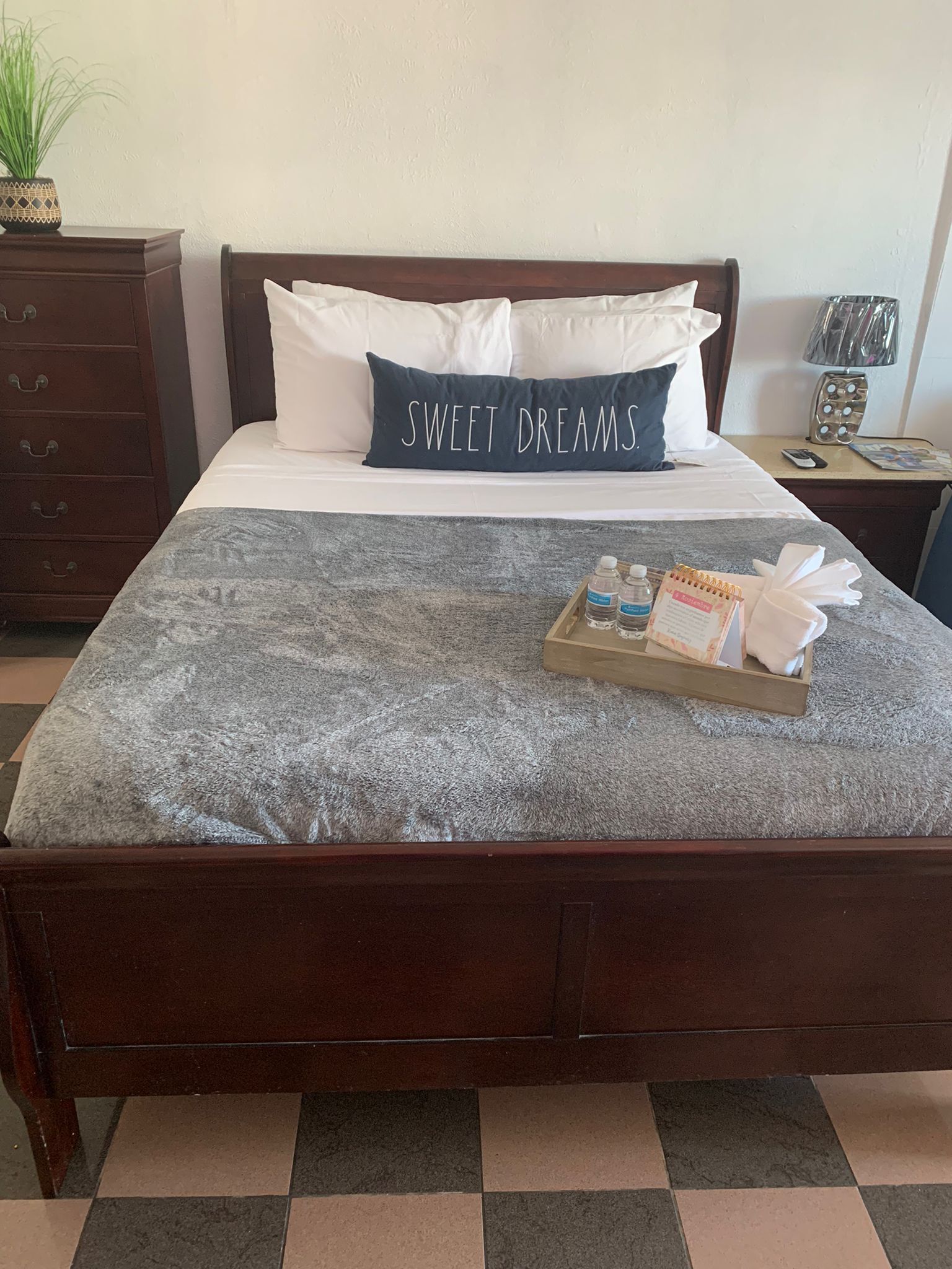 Un lit bien fait, recouvert d’une couverture grise à texture, un oreiller orné du mot « DOUX RÊVES », un plateau avec des articles de toilette et une table de chevet en bois avec une lampe.