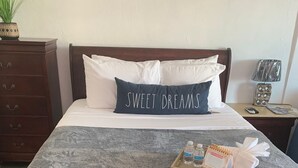 Ein ordentlich bezogenes Bett mit einer grauen, strukturierten Decke, einem Kissen mit der Aufschrift "SWEET DREAMS", einem Tablett mit Toilettenartikeln und einem Nachttisch aus Holz mit einer Lampe.