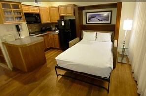 A compact hotel room with a bed, a small kitchenette, a microwave, a refrigerator, and a wooden cabinet with dishes.