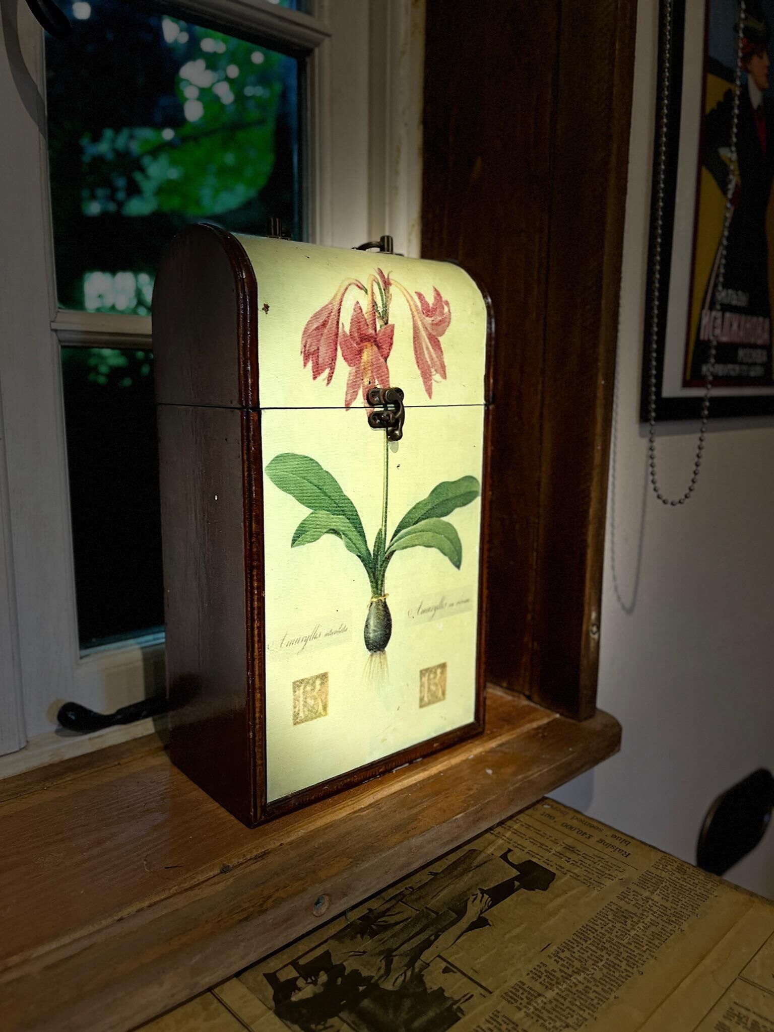 Een houten kist in vintage-stijl, versierd met bloemen en voorzien van een slot, staat op een houten oppervlak nabij een raam.