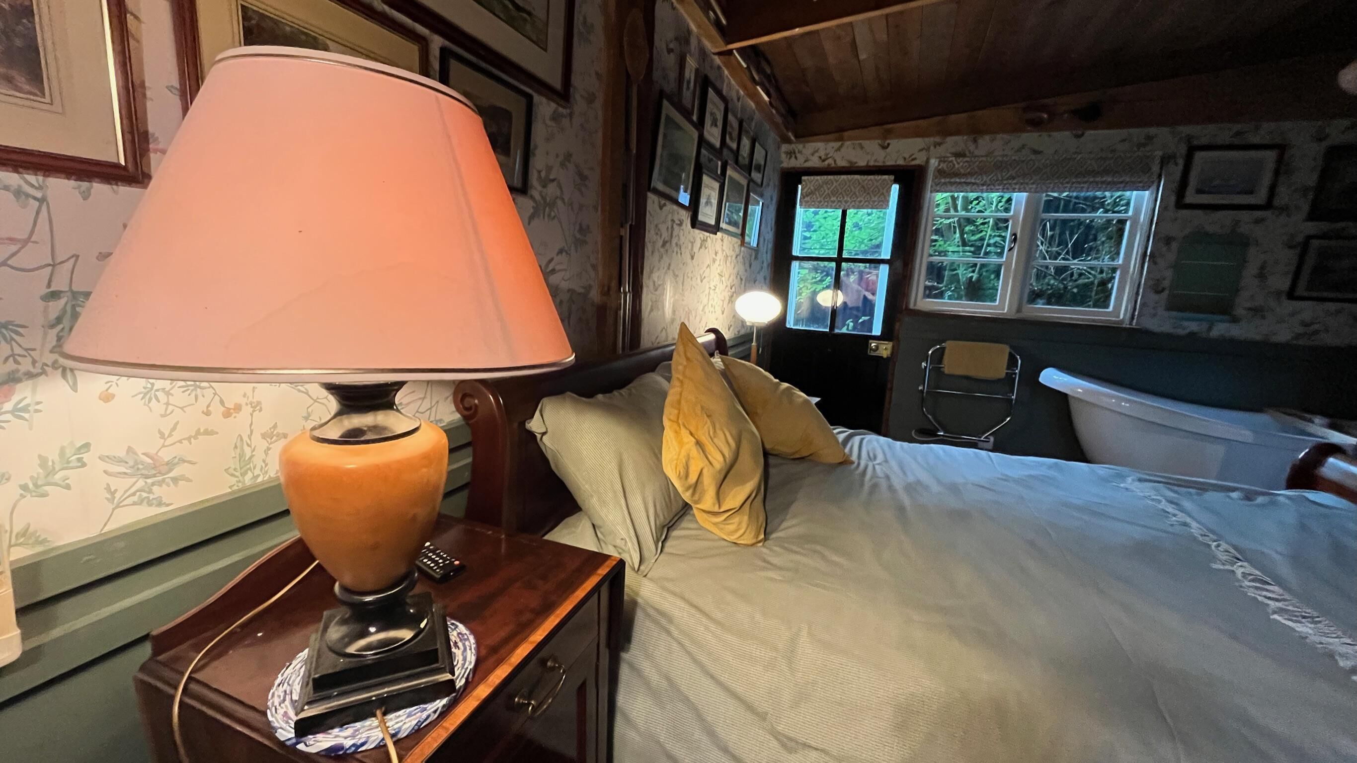 Ein Schlafzimmer mit Bett, Nachttisch mit Lampe, Fenster mit Blick auf Grünflächen und einem Badezimmer mit Badewanne.