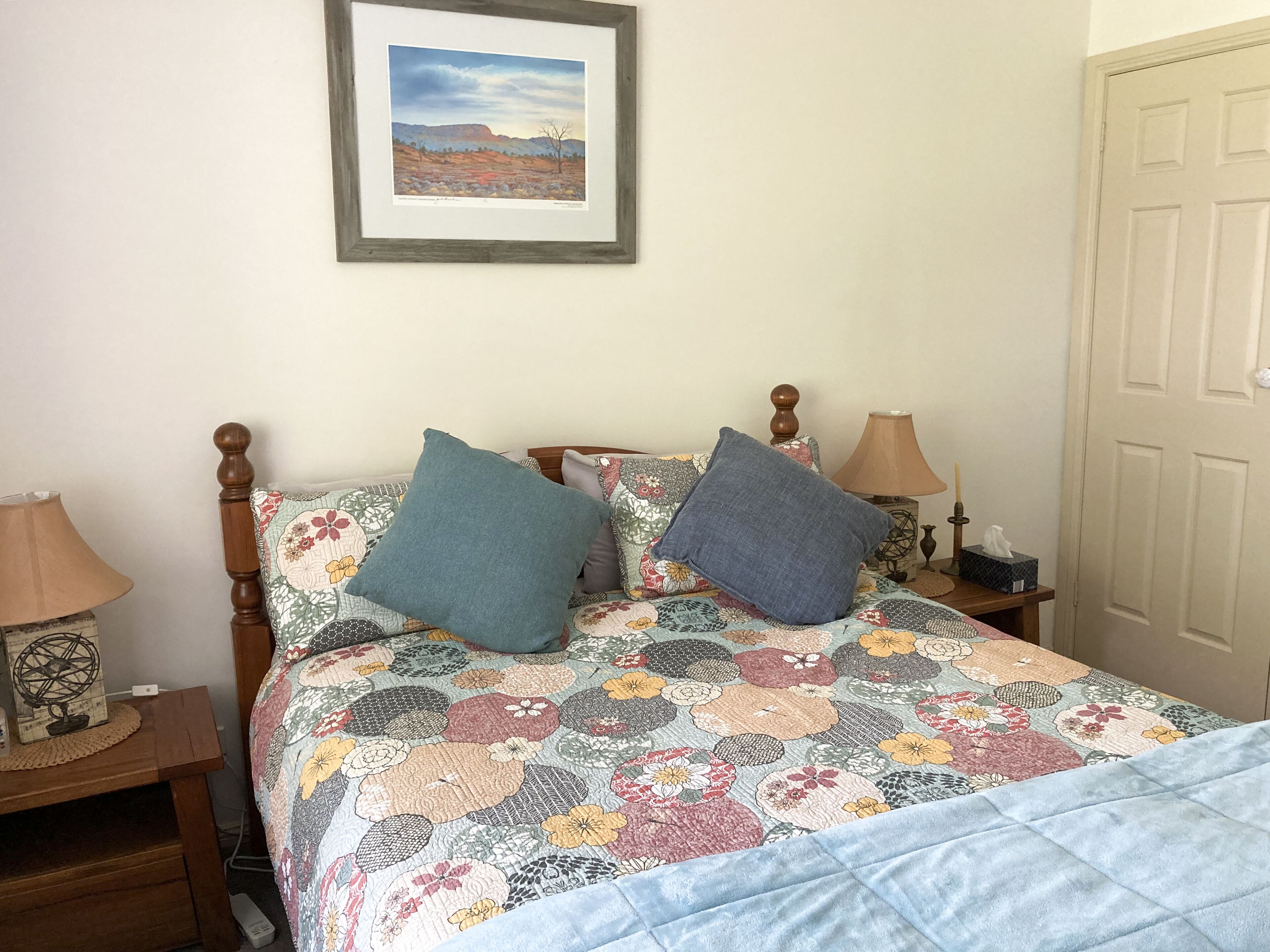 A bedroom with a floral-patterned bedspread, two pillows, a framed landscape picture on the wall, a bedside lamp, and a bedside table with a lamp and a tissue box.