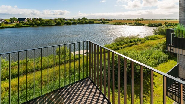 A balcony with a view of a river, greenery, and a distant building. - Apartment "Ankergold" with Dike View, Balcony & Wi-Fi (Nordstrand)