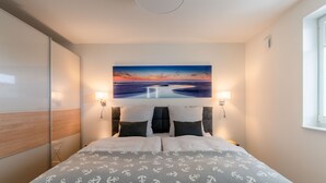 A hotel room with a bed, two bedside lamps, a painting of a sunset, and a window. - Apartment "Ankergold" with Dike View, Balcony & Wi-Fi (Nordstrand)