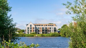 A modern multi-story building with a flat roof, large windows, and a mix of brick and wood facade, situated by a calm body of water. - Apartment "Ankergold" with Dike View, Balcony & Wi-Fi (Nordstrand)