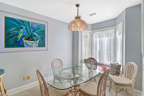 A dining area with a glass table surrounded by wicker chairs, a colorful pendant light, and a large framed painting on the wall.