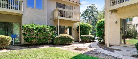 A residential building with a courtyard, featuring a covered patio, a small garden with manicured bushes, and a paved walkway.