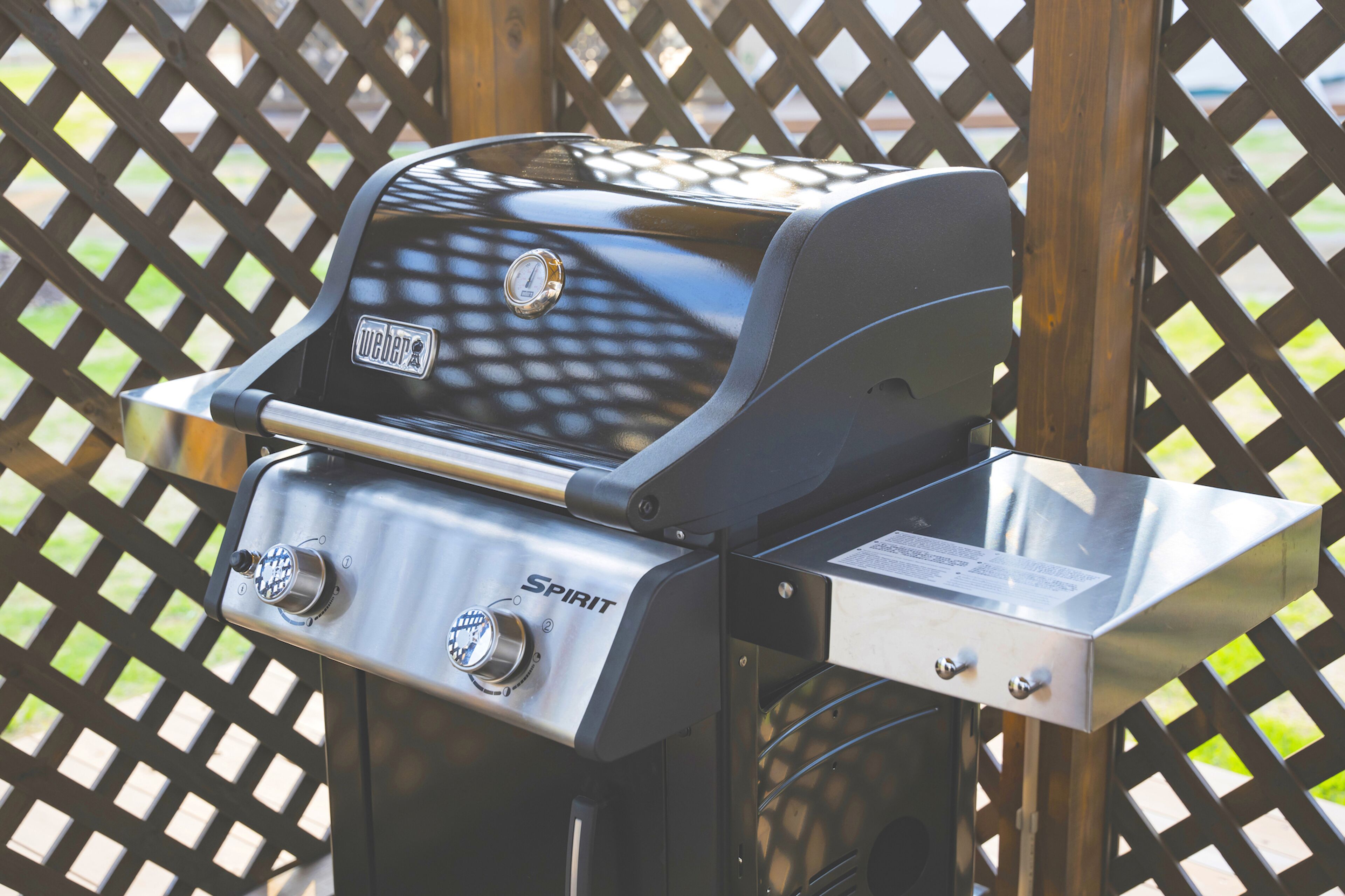 A Weber Spirit grill with a digital thermometer and control knobs, set against a wooden lattice background.