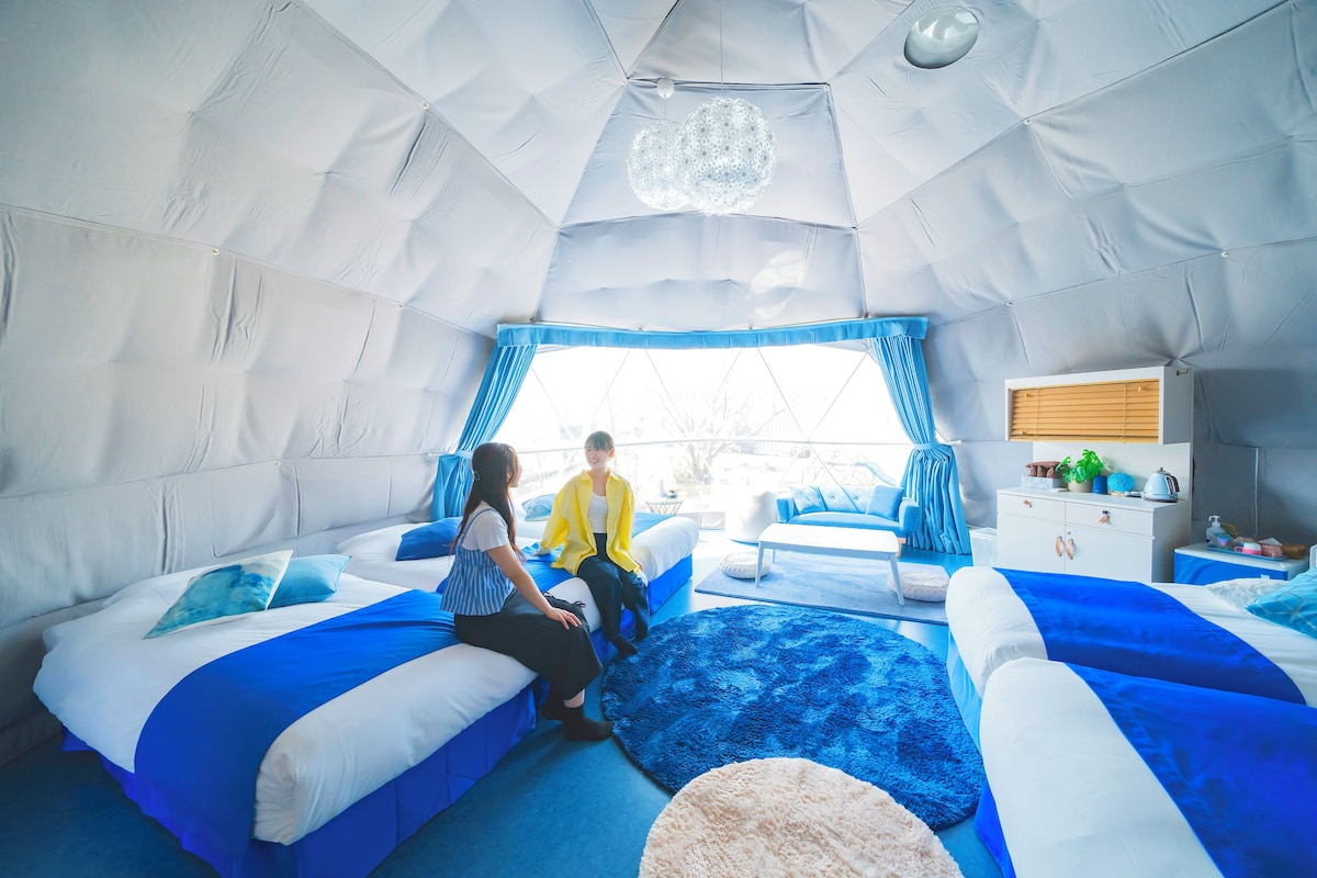 A cozy, tent-like room with two beds, a large window with blue curtains, and a white dresser with a mirror.