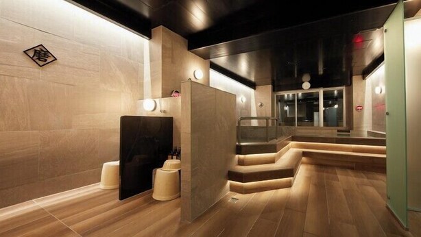 A modern spa with wooden flooring, a central seating area, and a glass elevator. - Rembrandt Cabin and Spa Shimbashi - Caters to Men (Tokyo)