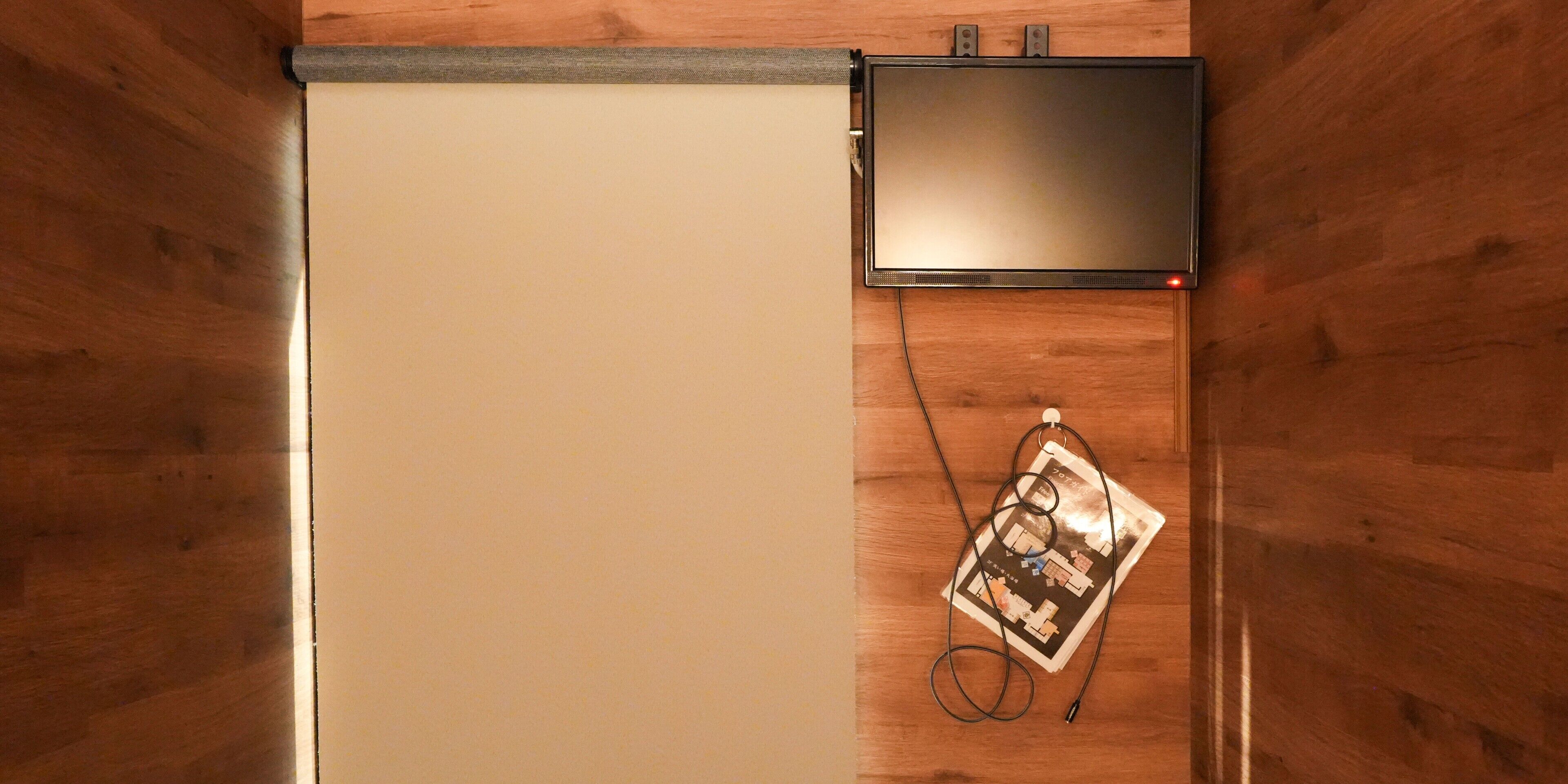 A room with a wooden floor, a closed door, a flat-screen TV mounted on the wall, and a map or diagram leaning against the wall.