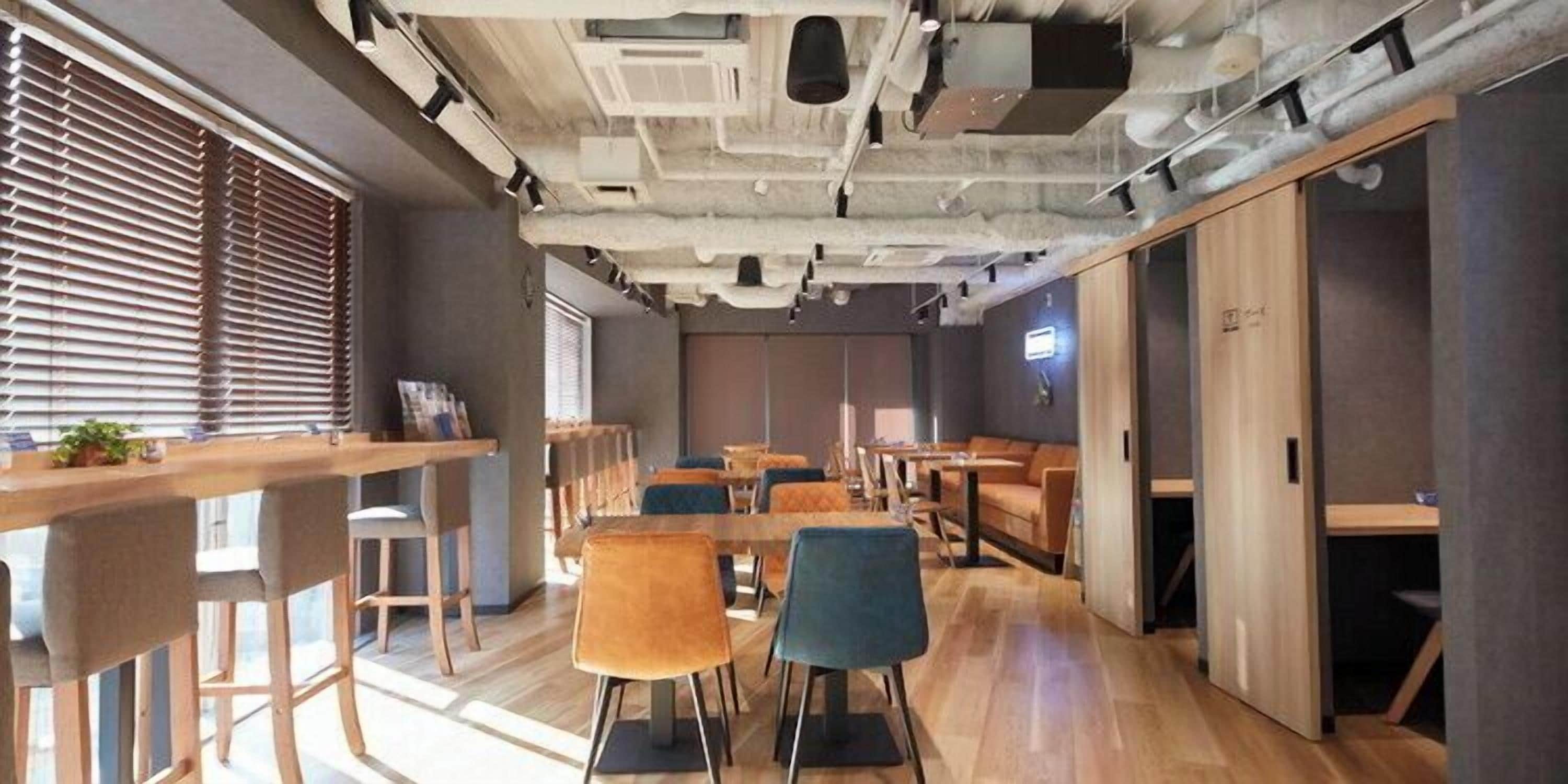 Un interior moderno con mesas y sillas de madera, techo alto con conductos expuestos y una variedad de espacios de asientos.
